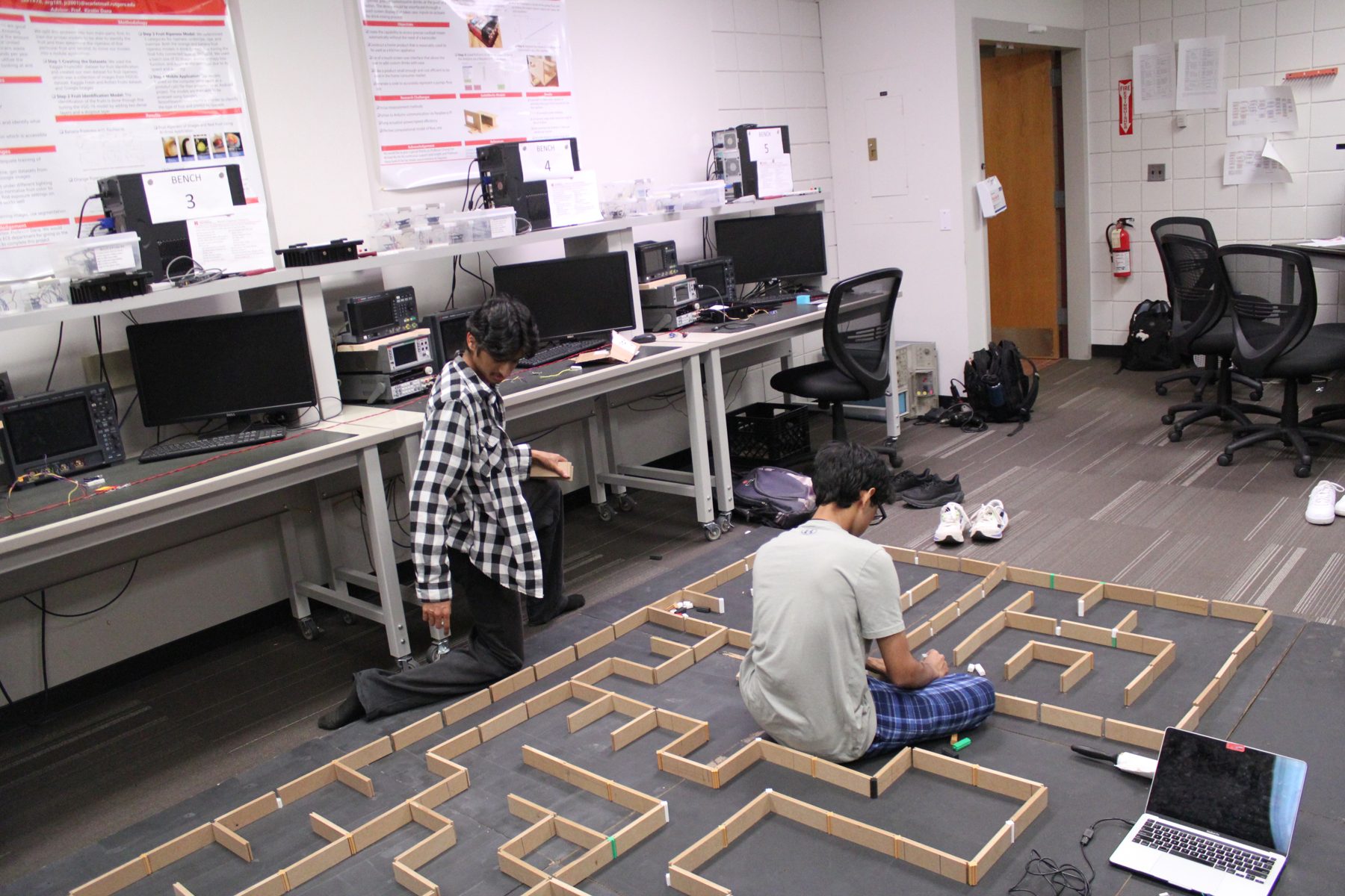 A practice maze of wooden walls laid out on the floor of a Rutgers engineering lab, two students working beside it