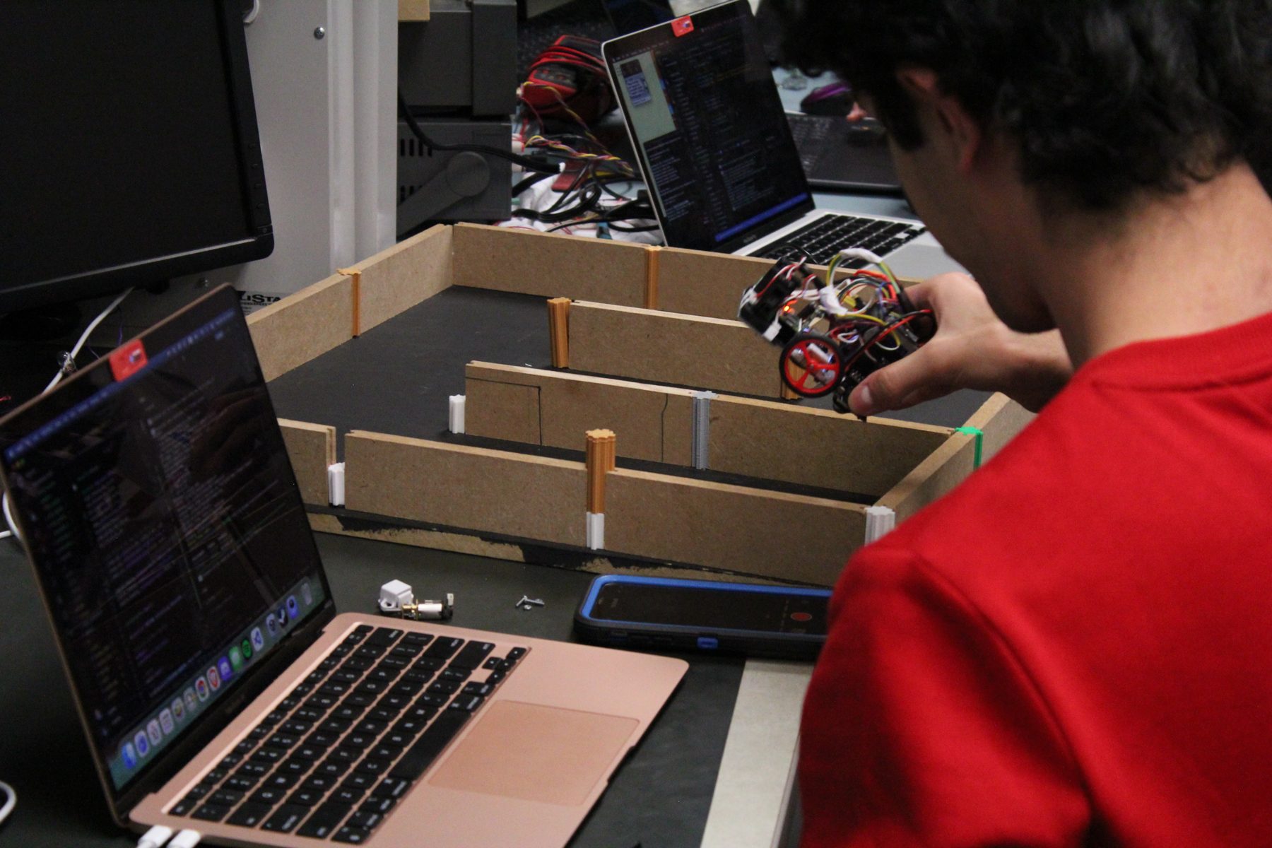 A teammate in a red shirt sets a robot down in a wooden practice maze, a laptop with code beside it