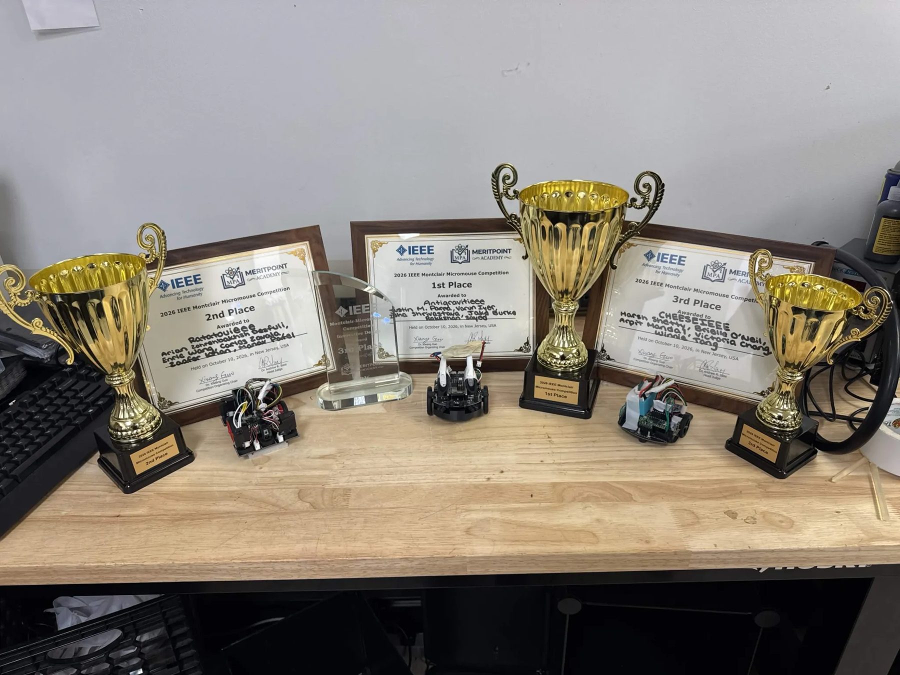Three trophies and three IEEE certificates on a lab bench, with the three winning robots between them